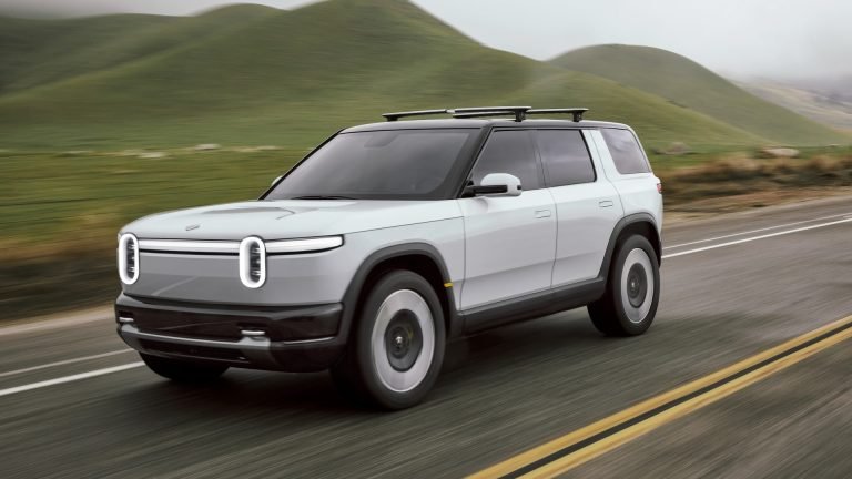 2024 Rivian R2 preview vehicle in light grey driving on a country road lined with hills, seen from the front three-quarter