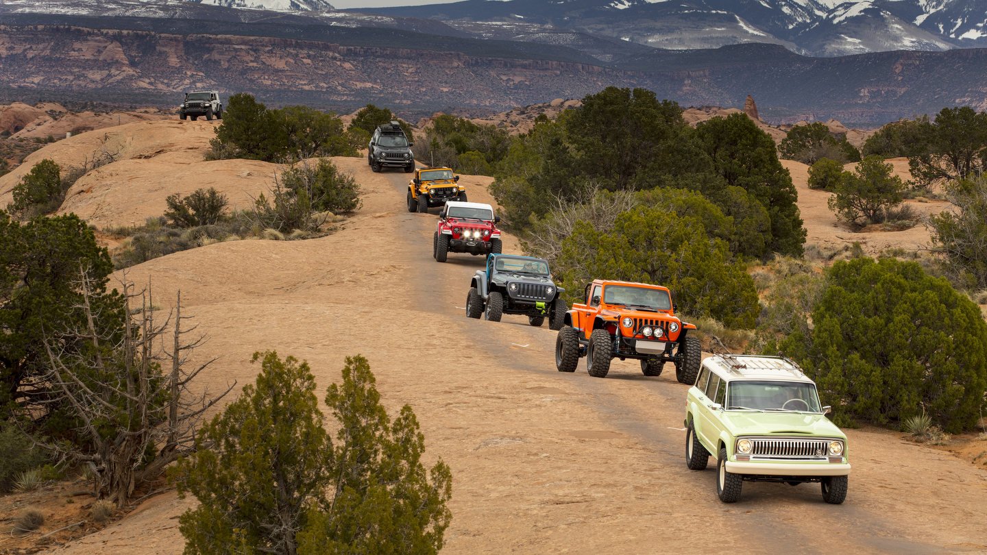 Line of Jeep vehicles, led by a vintage green Wagoneer, driving across sandstone slickrock near Moab, Utah, with snowcapped mountains behind.