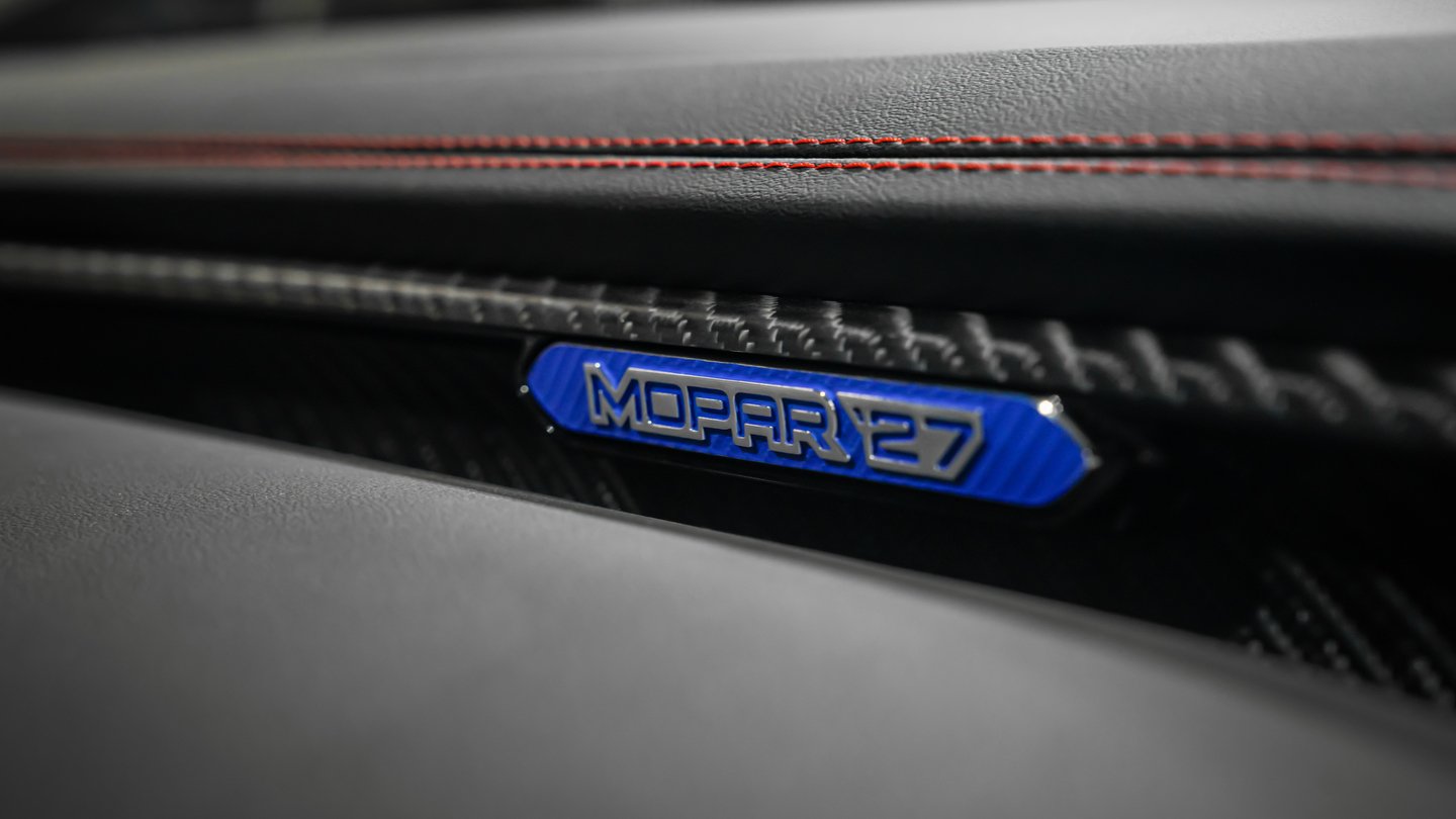 Mopar ‘27 Badge, SEMA 2026 Teaser Blue Mopar ‘27 badge on a black dashboard with carbon-fibre-pattern trim and red contrast stitching