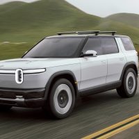 2024 Rivian R2 preview vehicle in light grey driving on a country road lined with hills, seen from the front three-quarter